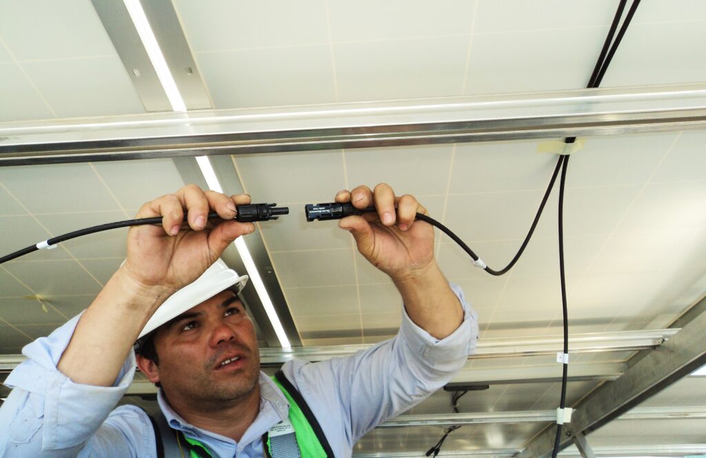 An electrician setting up a wiring connection. He has a hard-hat and high visibility vest on as protective equipment..